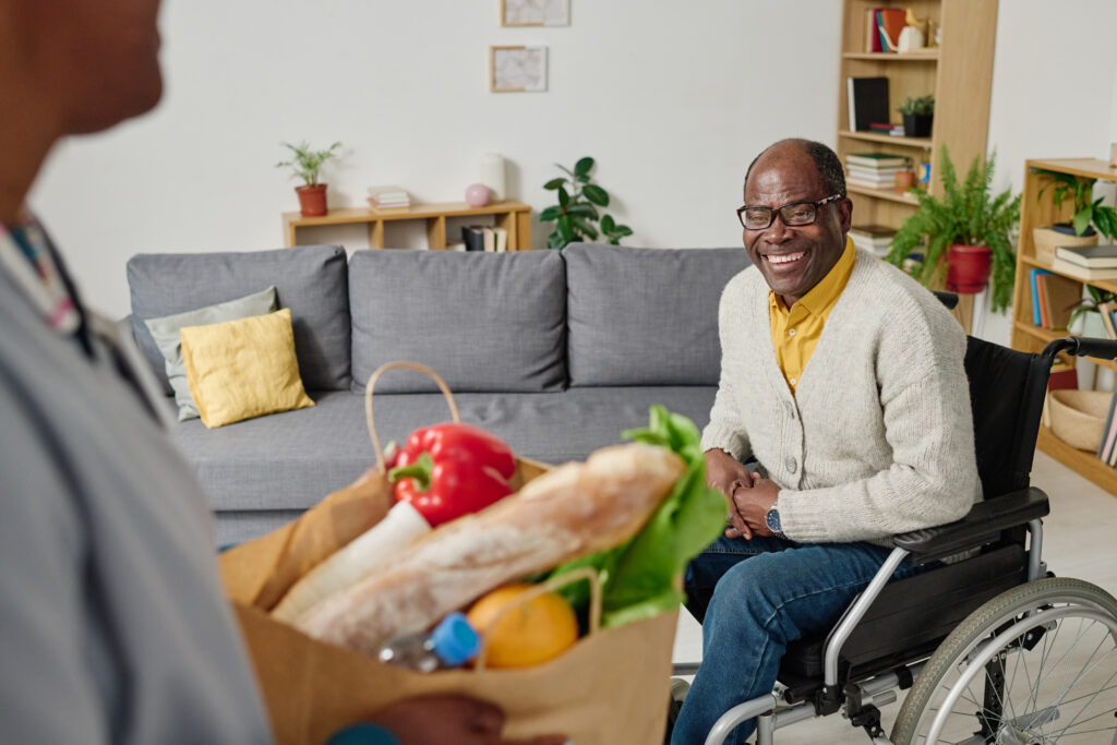 African happy man with disability sitting in wheelchair and meeting caregiver bringing groceries for him